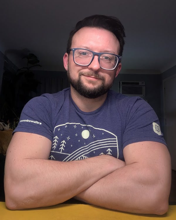 Justin Sovine in glasses and a navy t-shirt, smiling, arms crossed on a table
