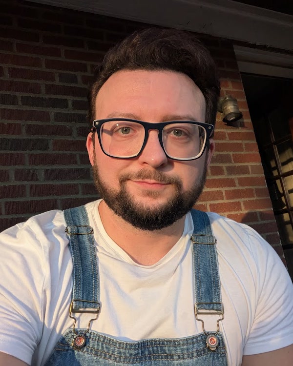 Justin Sovine in glasses, a white t-shirt and denim overalls, in front of a brick wall in evening sun